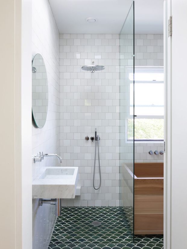 Bright, compact contemporary bathroom viewed through an open doorway. The room features white rectangular wall tiles, a green and white scallop-pattern tiled shower floor, a frameless glass shower screen, wall-mounted shower fittings, a white stone or ceramic basin, an oval wall mirror, and a timber vanity beneath a window. The palette is predominantly white with green and warm timber accents.