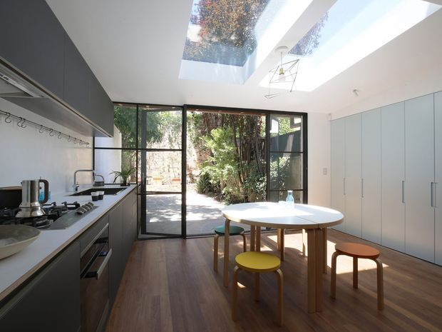 Bright contemporary kitchen-dining space with extensive dark built-in cabinetry and open shelving on the left, a large white island bench, and a round white dining table with timber stools and chairs. Full-height black-framed glazed doors open to a leafy courtyard, while large skylights bring in abundant daylight. Warm timber flooring contrasts with the white walls and dark joinery.