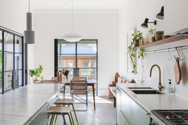 Bright, refined kitchen with a long pale stone island, integrated seating, white cabinetry, open timber shelving, brass tapware, black-framed glazing and pendant lighting. A dining area and living space are visible beyond, with abundant natural light and indoor plants adding a fresh, contemporary feel.
