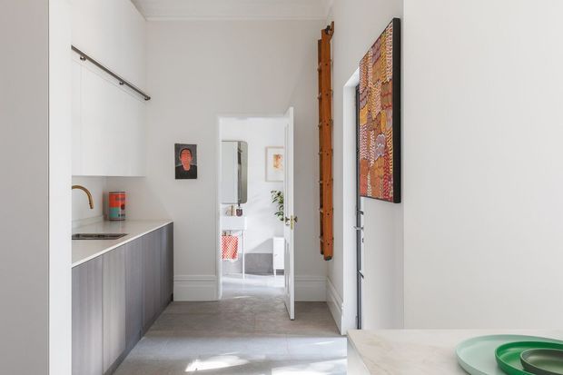 A bright, narrow galley kitchen viewed toward an open doorway and adjoining space. The kitchen has white upper cabinetry, dark vertically grained lower cabinetry, a slim stainless-steel rail, a pale benchtop, and light grey tiled flooring. A tall timber feature panel and colourful wall artwork add warmth, while a small portion of a dining or work surface with green trays is visible in the foreground.
