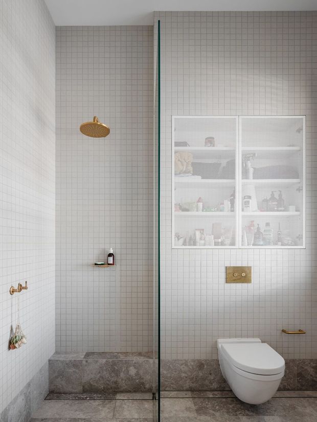 Minimalist contemporary bathroom with full-height small square white wall tiles, a grey stone tiled floor and shower base, and a frameless glass shower screen. The shower has a brushed brass rain showerhead and controls, while a wall-hung white toilet sits beneath a recessed glass-fronted storage cabinet. Brass plumbing fixtures and hardware provide warm accents.