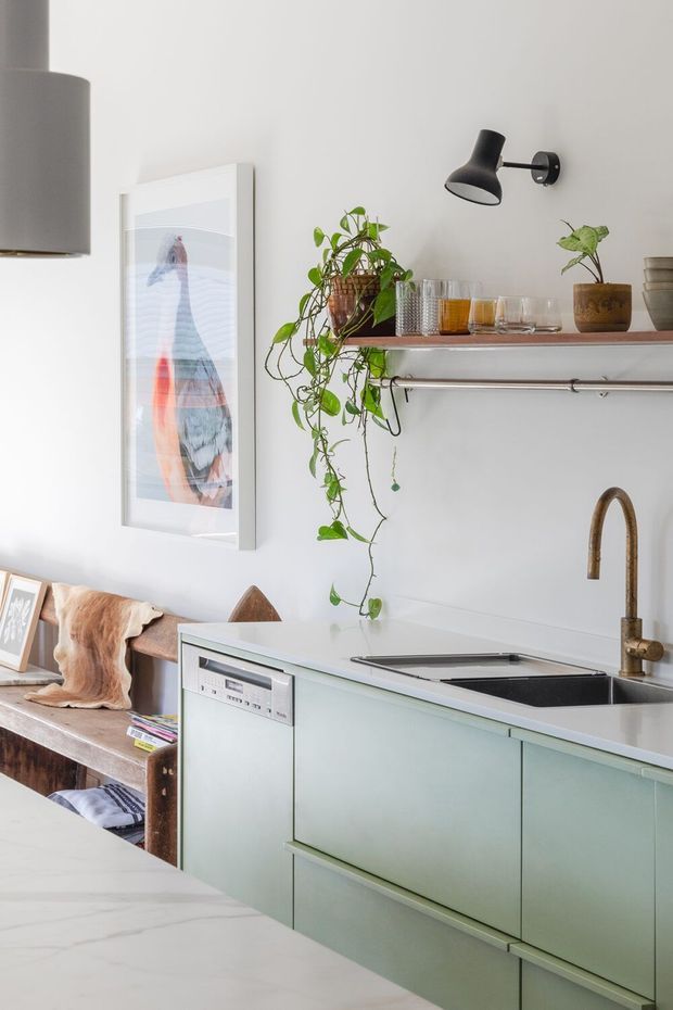 Bright, compact contemporary kitchen with pale green fixed cabinetry, a white stone-look benchtop, integrated dishwasher, black cooktop, and a brass-toned gooseneck tap. Open timber shelving displays glassware and potted trailing plants, with a black wall light and framed artwork above a timber dining bench. A marble-look island or foreground worktop occupies the lower left.