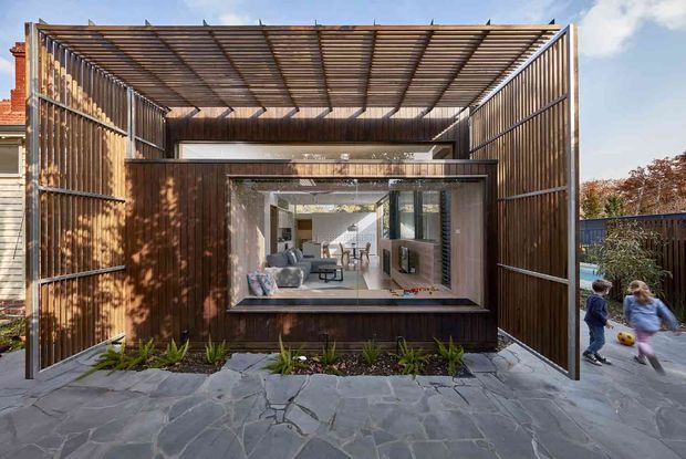 Exterior view of a contemporary residential extension with a large timber-clad pavilion or outdoor room framed by a deep open pergola and vertical slatted screens. The projecting glazed opening reveals a bright living area inside, while irregular stone paving and planted beds lead toward the entrance. The design uses strong indoor-outdoor connections, layered screens, and a sheltered courtyard-like setting.