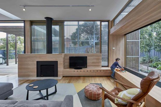 A bright contemporary living room with extensive floor-to-ceiling glazing, vertical screening, warm timber wall panelling, and a built-in fireplace and television unit. The room contains a grey sectional sofa, round dark coffee table, leather lounge chair, ottoman, and a low window-side bench. A child is present near the window, and greenery is visible outside.
