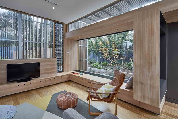 A bright, contemporary living room with extensive timber built-in joinery, including a low media console, window seat, and a large timber-framed opening to the garden. Vertical slatted screening and clerestory-style glazing provide filtered natural light, while a mid-century-style lounge chair, patterned rug, ottoman, and television furnish the space.