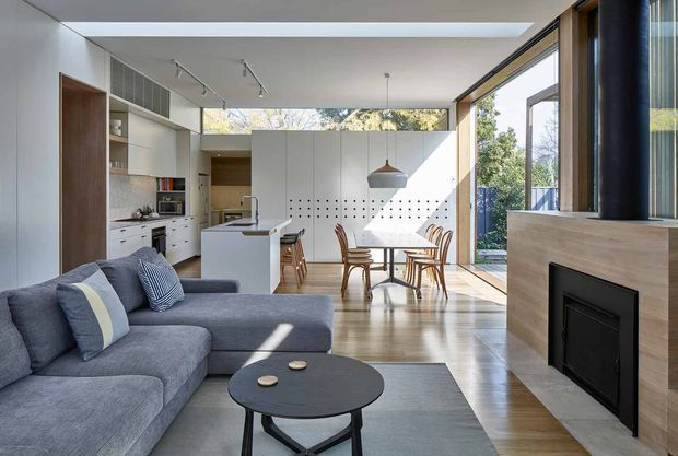 Bright open-plan contemporary living space combining a lounge, kitchen and dining area. A large grey sectional sofa and round black coffee table occupy the foreground, with a compact white island and timber dining table beyond. Full-height glazing opens the room to greenery, while a timber-clad fireplace wall anchors the right side. White cabinetry, pale timber flooring, a perforated feature wall and a sculptural pendant complete the clean, sunlit interior.
