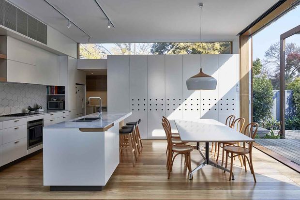 A bright open-plan kitchen and dining area with white flat-panel cabinetry, a large island with seating, integrated appliances, and a white dining table surrounded by timber chairs. Warm timber flooring, a sculptural pendant light, track lighting, and full-height glazing connect the room to a timber deck and leafy outdoor setting. The clean-lined interior receives strong natural light and has a contemporary minimalist feel.