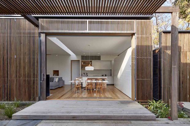 A timber-clad contemporary pavilion or extension viewed from an outdoor deck, with a large fully opened glazed wall revealing a combined kitchen and dining area. The interior features white walls and cabinetry, warm timber flooring, a dining table with bentwood chairs, a pendant light, and a partially visible lounge. Vertical timber screening and a slatted pergola create a strong layered facade and shaded outdoor threshold.