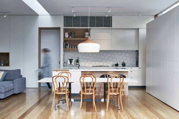 Bright open-plan kitchen and dining area with white built-in cabinetry, a patterned hexagonal tile backsplash, central island, and a timber dining table surrounded by bentwood chairs. Light timber flooring, a blue sofa, integrated storage, skylight-like ceiling openings, and contemporary pendant and track lighting create an airy, clean interior.