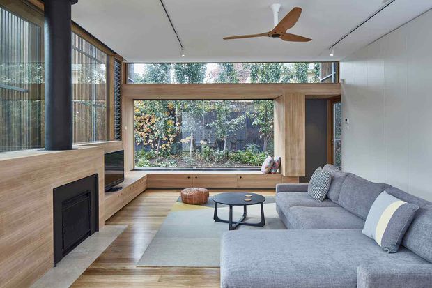 Bright contemporary living room with a large grey sectional sofa, low round coffee table, built-in timber window seat and cabinetry, and a timber-clad fireplace with black flue. Floor-to-ceiling glazing and clerestory windows overlook a leafy courtyard, while a timber ceiling fan and recessed linear lighting complete the calm, natural interior.
