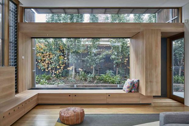 A warm, contemporary living room centred on a large garden-facing window. Built-in timber cabinetry and a window seat frame the glazing, with a timber-lined overhead beam, clerestory glazing, and a glazed door at right. The room includes a low upholstered ottoman, a partial sofa, a patterned cushion, and a muted area rug. A planted courtyard is visible beyond the windows.