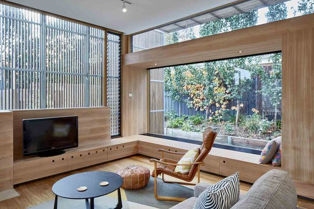 A warm, contemporary living room with extensive floor-to-ceiling glazing, timber wall panelling and built-in window seating. The room includes a low timber media console, wall-mounted television, round dark coffee table, upholstered sofa, leather-and-timber lounge chair and ottoman. Vertical exterior screening and a planted courtyard are visible through the windows, creating a calm connection to the outdoors.