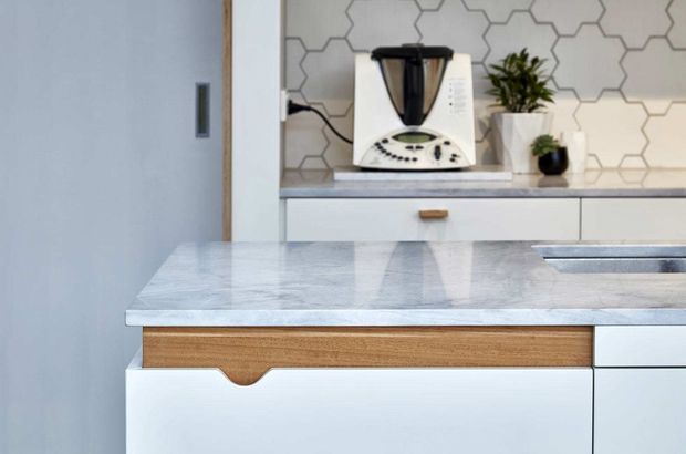 Close-up view of a contemporary kitchen featuring a pale stone island benchtop, white cabinetry with integrated wood drawer fronts and finger-pull details, and a geometric hexagonal tiled splashback. Small appliances and plants sit on the rear counter.