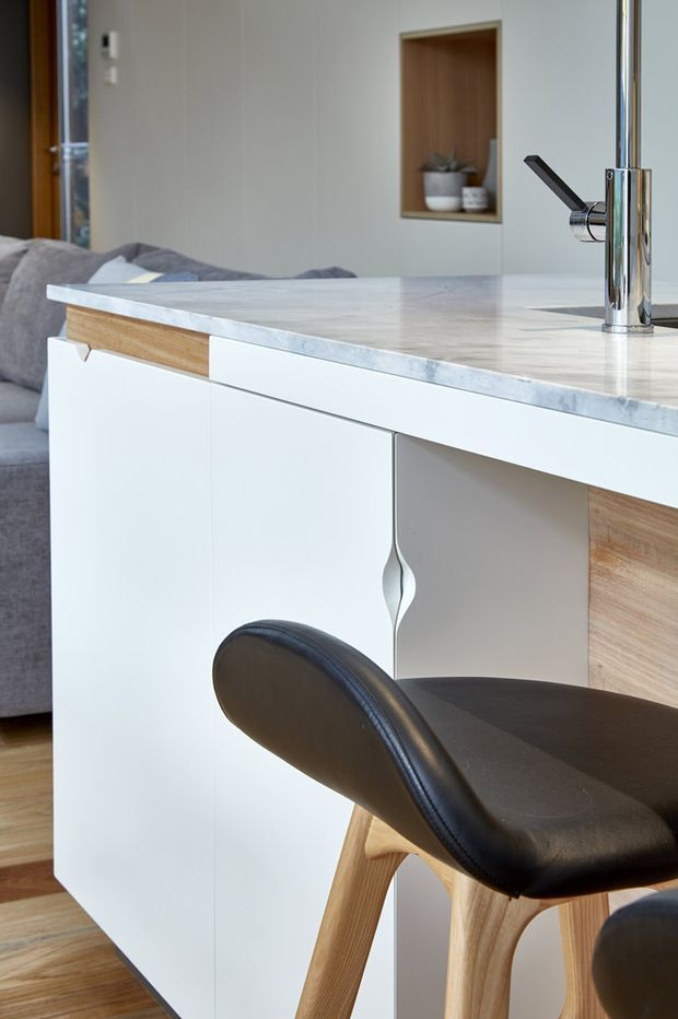 Close-up interior view of a contemporary kitchen island adjoining a living area. The island has white flat-panel cabinetry with a distinctive sculpted vertical handle, warm timber drawer fronts and side panels, and a pale veined stone benchtop with an integrated chrome mixer tap. Black leather and timber bar stools sit at the island, with a grey sofa and a recessed timber-lined display niche visible behind.