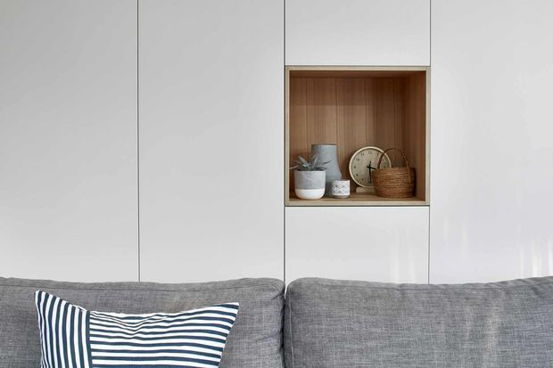 Close-up interior detail of a contemporary built-in white wall unit with a recessed open display niche lined in warm timber. The niche contains ceramics, a clock and a woven basket, while the foreground shows a grey upholstered sofa with a navy-and-white striped cushion.