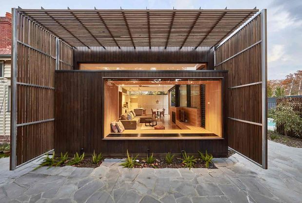 Exterior view of a compact contemporary pavilion or extension clad in dark vertical timber, with a large open steel-framed pergola-like screen enclosing the facade. A broad picture window reveals a warmly lit living area and dining space inside. The foreground has irregular stone paving and a narrow planted garden bed.