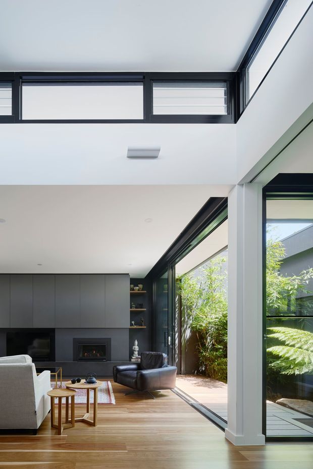 A contemporary open-plan living room with a double-height ceiling and clerestory windows. The room features dark built-in cabinetry, a black fireplace wall, timber flooring, expansive black-framed glazing, and a compact timber deck with planted greenery outside. A white sofa, dark leather armchair, and small timber tables create a warm, modern seating area.