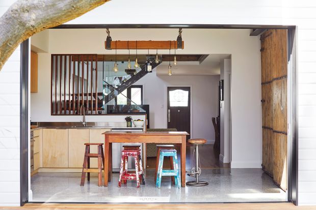 Bright open-plan kitchen and dining area viewed through a large framed opening, featuring a timber island bench, pale timber cabinetry, stools, pendant lighting, exposed timber beams, a dark timber staircase with vertical slat screen, and a substantial timber sliding door. Polished concrete flooring and white walls create a clean, contemporary warehouse-inspired interior.