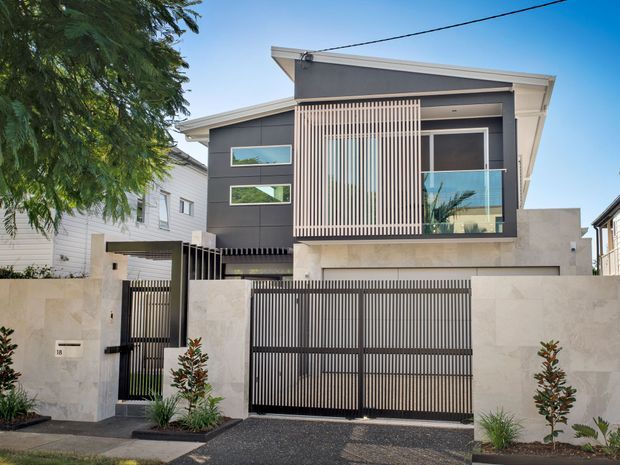 Street-facing view of a contemporary multi-storey urban residence with a flat, asymmetrical roofline, dark grey rendered upper facade, pale rendered lower walls, extensive vertical screening, glazed balcony balustrade, and a large slatted driveway gate. The property is framed by neighbouring homes, a paved driveway, low landscaping, and a boundary wall.