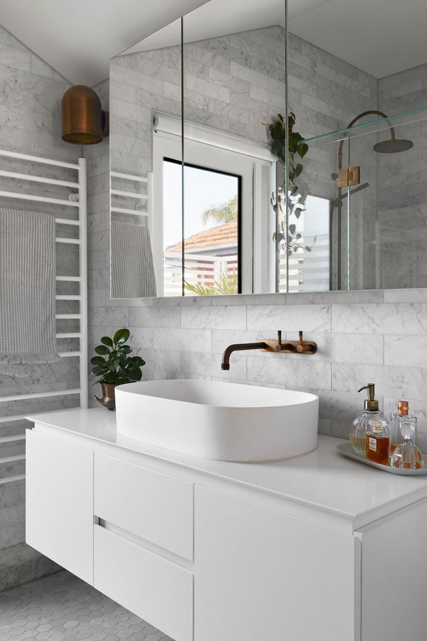 A bright, refined bathroom featuring a white freestanding vessel basin on a wall-mounted white vanity, grey-white marble-look wall tiles, a small hexagonal tile floor, a large mirrored cabinet, a white heated towel rail, and a brushed bronze wall-mounted tap. A glass shower enclosure is visible to the right.