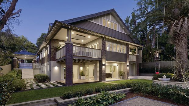 Exterior dusk view of a substantial two-storey suburban residence with a steep gabled roof, dark horizontal cladding, brick columns, broad covered balconies, and extensive glazed openings. The landscaped front garden includes hedges, paving, planting, and a small water feature or planter area.