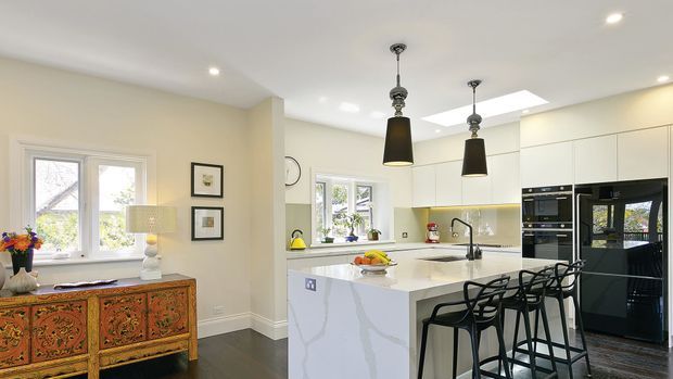 Bright open-plan kitchen with a large white waterfall island, black bar stools, integrated black appliances, pale cabinetry, pendant lights, skylight, and dark timber flooring. A decorative sideboard and windows are visible in the adjoining living area.