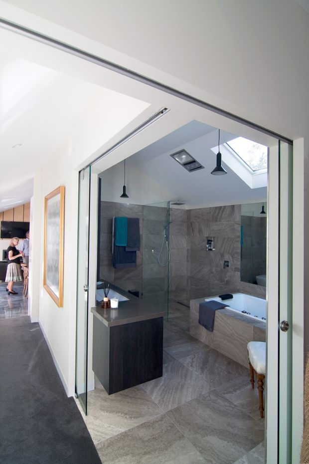 A contemporary bathroom viewed through wide frameless glass doors from a hallway. The room features large grey stone-look tiles across the floor and walls, a freestanding or partially enclosed bathtub, a glass walk-in shower, dark wood-toned vanity cabinetry, a skylight, recessed ceiling lighting, and black pendant lights. A framed artwork and adjacent interior space are visible beyond the bathroom.