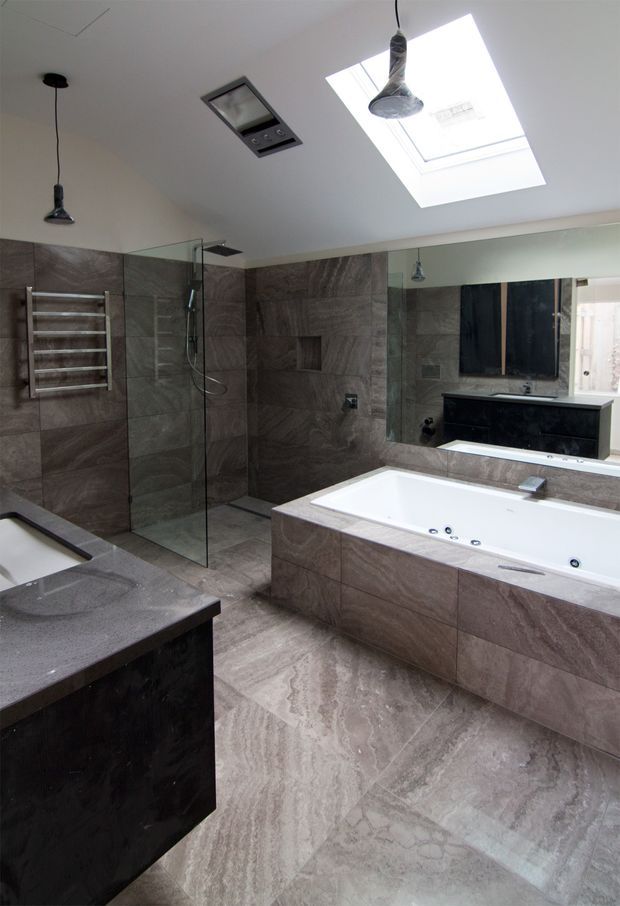 A spacious contemporary bathroom featuring a large freestanding-style built-in bathtub, frameless glass shower enclosure, double vanity with dark cabinetry and stone benchtop, extensive grey-brown natural stone tiling, a heated towel rail, wall-to-wall mirror, recessed ceiling lighting, pendant lights, and a large skylight.