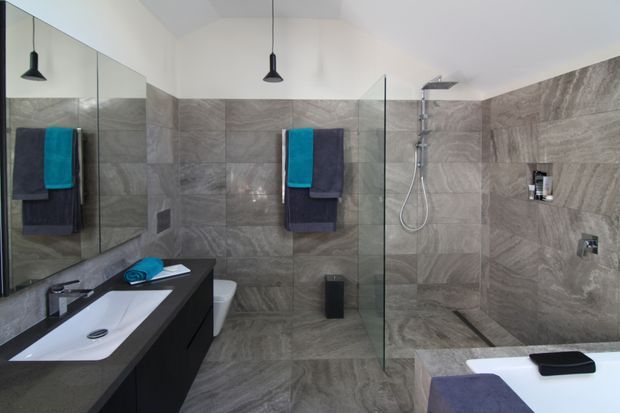 Contemporary bathroom with large-format grey stone-look tiles covering the walls and floor, a frameless glass walk-in shower, wall-mounted rain shower and handheld fixture, black floating vanity with white integrated basin, mirrored wall panels, and dark pendant lighting. Bright, clean, and refined with strong grey and black contrast.