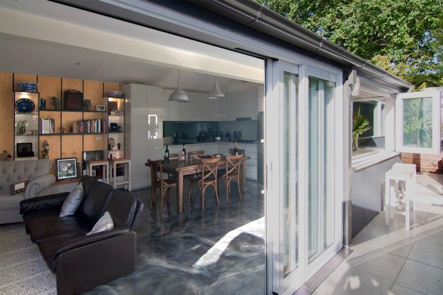 A bright contemporary open-plan living and dining area viewed through wide open glazed folding doors. The interior features a dark leather sofa, built-in timber display shelving, a timber dining table with cross-back chairs, pendant lights, and dark polished flooring. White cabinetry and walls create a clean contrast with the timber joinery. A paved outdoor terrace and additional glazed extension are visible beyond the doors.