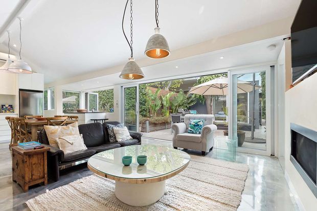 Bright open-plan living room with a black sofa, armchair, round marble-topped coffee table, textured rug, timber side cabinet, pendant lights, and a wall-mounted fireplace. Full-height windows and glazed doors open the room visually to a leafy outdoor courtyard or patio.