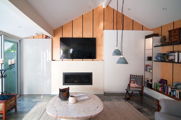 Bright contemporary living room featuring a white built-in media and fireplace wall, a large wall-mounted television, warm vertical timber wall panelling, integrated white cabinetry, open shelving, pendant lights, and a round stone coffee table over a textured rug. Glazing at left brings in natural light and reveals greenery outside.
