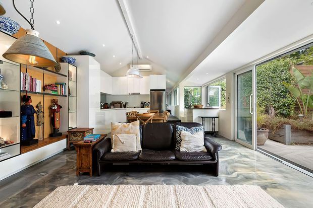 Bright, elongated open-plan living space with a black leather sofa in the foreground, integrated display shelving along the left wall, and a compact white kitchen with island and timber stools at the rear. Full-height glazed sliding doors open to a planted courtyard, while large pendant lights, polished concrete-style flooring, and a white angled ceiling create an airy contemporary interior.