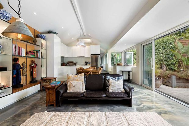 Bright open-plan living room with a black leather sofa in the foreground, a compact white kitchen and dining area beyond, extensive sliding glass doors opening to a planted courtyard, and built-in display shelving along the side wall. The space combines polished concrete flooring, white cabinetry and walls, timber accents, and eclectic decorative objects with abundant natural light.