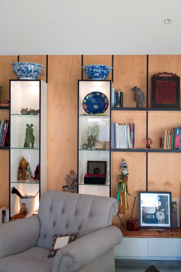 A styled living room with a large built-in timber display and shelving wall. The joinery combines warm wood panels with dark framing, illuminated glass display niches, open bookshelves, and a low white cabinet. The shelves hold ceramics, sculptures, books, textiles, and framed artwork. A large tufted grey upholstered armchair occupies the foreground, creating a layered, eclectic and curated interior.