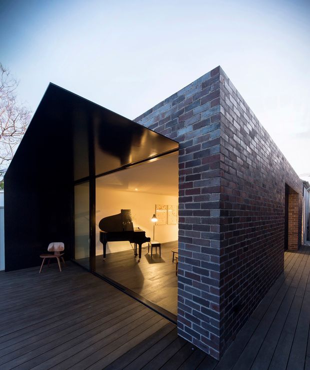 Exterior view of a contemporary, sharply geometric building with a dark metal-clad angular volume adjoining a tall exposed brick masonry wall. Large glazed openings reveal a warmly lit interior with a grand piano, while a broad dark timber deck wraps around the structure.