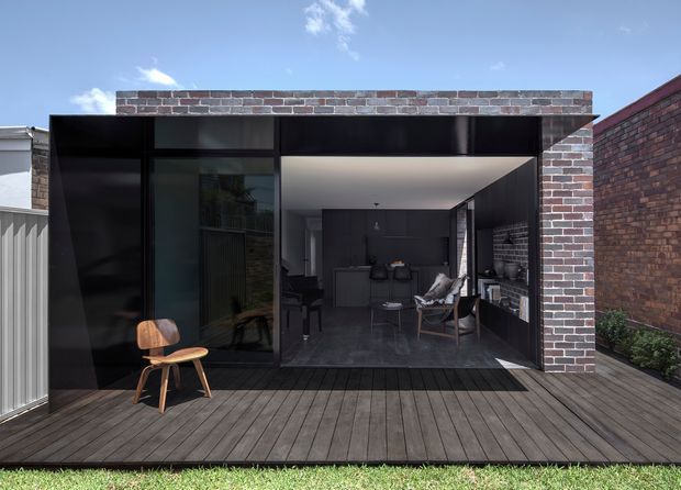 A compact contemporary backyard extension or pavilion opens onto a broad timber deck through large dark-framed glazed doors. The exterior is framed in exposed red-brown brick with black metal panels and glazing, while the interior contains a dark minimalist kitchen and lounge area with a dining table, chairs, and built-in cabinetry. A neighbouring brick wall and small garden edge are visible around the deck.