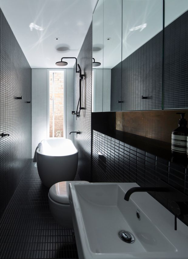 A narrow, high-contrast contemporary bathroom with dark small-format tiled walls and floor, a freestanding white bathtub, wall-mounted shower fittings, toilet, and a white rectangular basin with matte black tapware. A large mirrored cabinet reflects the shower and tiled surfaces, while a slim window brings in daylight. The palette is predominantly black, white, and charcoal with a warm metallic-toned backsplash niche.