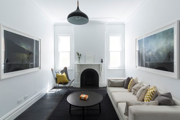 Bright, compact formal living room with white walls, traditional ceiling cornices and skirting, a central black pendant light, a white mantel fireplace, dark timber flooring, and a large light-coloured sectional sofa. Two large framed artworks flank the fireplace, with a black round coffee table and a yellow accent chair adding contrast.
