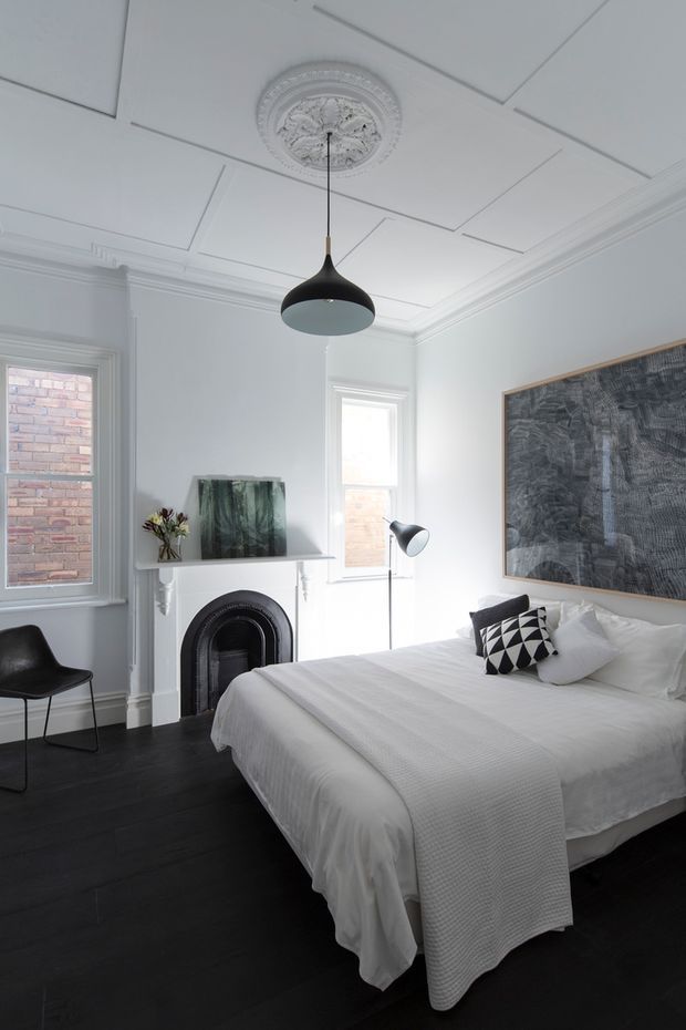 A refined bedroom with a large white upholstered bed dressed in white linen, set against a white decorative interior. The room features a black-painted timber floor, ornate ceiling medallion and cornices, wall panel mouldings, a dark arched fireplace, black pendant and floor lamps, and monochrome artwork and cushions. Tall windows reveal brickwork outside, while a simple black chair occupies the corner.