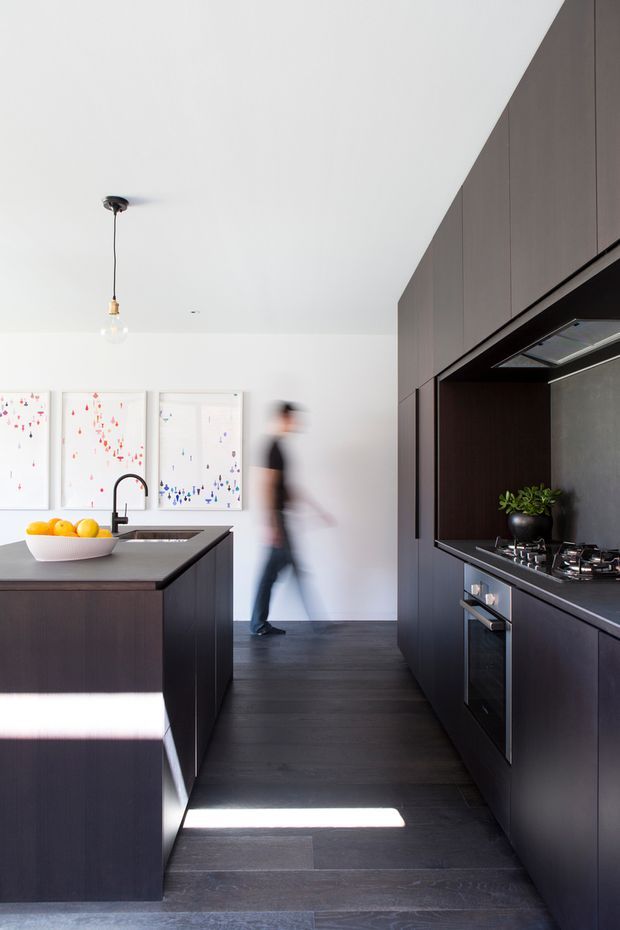 A contemporary galley-style kitchen with a central island, dark timber cabinetry, dark flooring, pale walls, and integrated appliances. The island includes a sink with a black gooseneck tap and a dark benchtop, while the opposite wall contains a cooktop, oven, rangehood, and additional cabinetry. Minimal artwork and a bowl of fruit provide limited decorative accents, with strong sunlight creating bright bands across the floor and island.