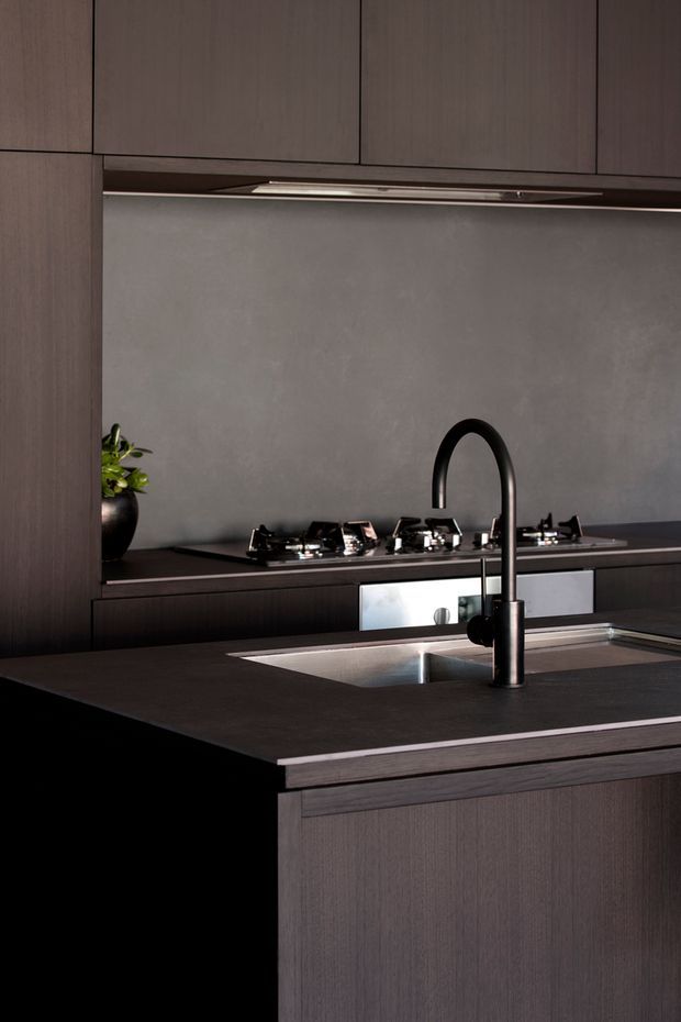 Close-up of a sophisticated dark-toned kitchen with flat-panel timber-look cabinetry, a large dark benchtop island, integrated stainless-steel sink, black gooseneck tap, gas cooktop, and a grey textured splashback. The composition emphasizes the fixed joinery, island and cooking zone in a moody, minimalist interior.