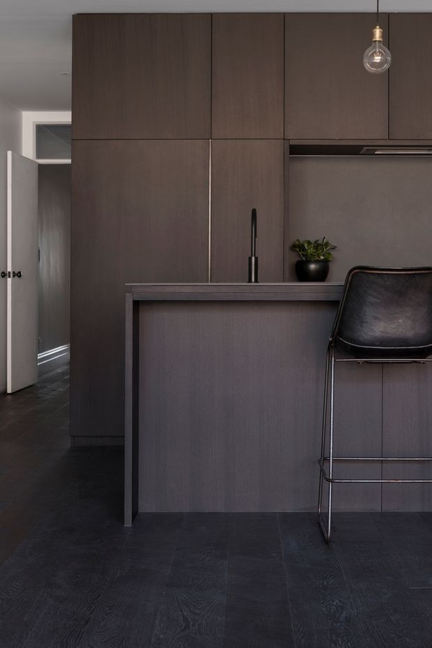 A moody, compact contemporary kitchen viewed from the living area, with full-height dark timber cabinetry, a matching island or peninsula, integrated rangehood, black tapware, and dark flooring. A black leather bar stool and exposed pendant bulb add contrast, while a hallway is visible at left.