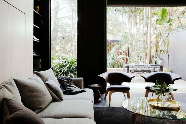 A refined contemporary living room with a large light-grey sectional sofa, two sculptural dark lounge chairs, small timber stools, and a round glass coffee table. Full-height black-framed glazing opens the room visually to a lush planted outdoor area, while pale built-in cabinetry lines the left wall. The interior has a calm, sophisticated palette with strong contrast between light upholstery, dark frames and furniture, and abundant greenery outside.
