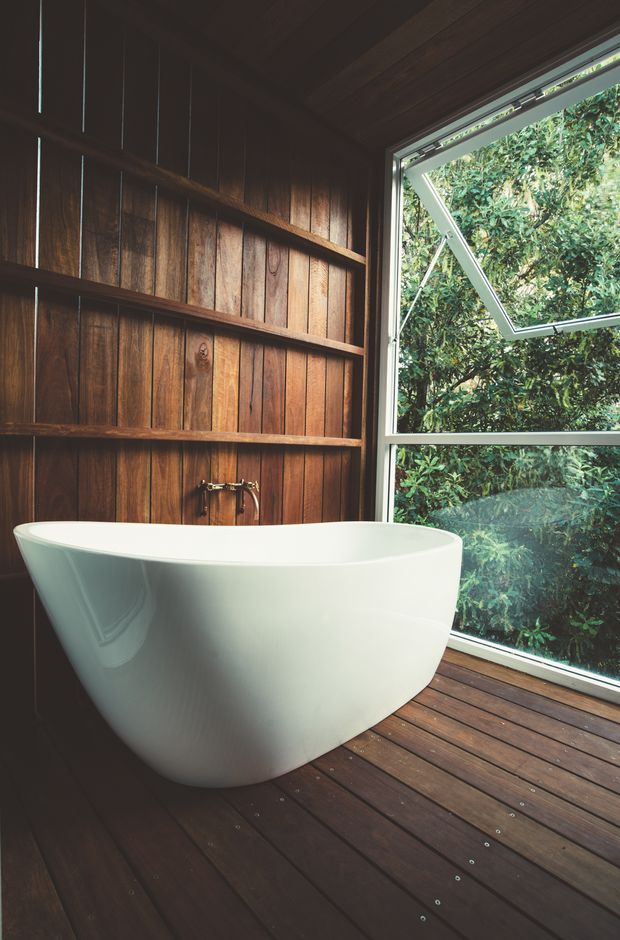 A compact, dark timber-lined bathroom featuring a sculptural white freestanding bathtub, wall-mounted brass tapware, and a large aluminium-framed window with an awning section opening toward dense greenery. The bathroom floor is finished in dark timber decking, creating a warm, nature-connected atmosphere.