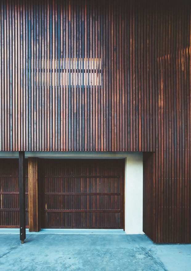 A close-up architectural view of a dark timber-slatted facade, featuring dense vertical battens, a recessed timber garage or entry door, and a small horizontal opening with light visible behind it. The lower foreground is a smooth blue-grey concrete surface. The composition emphasizes rhythm, texture, shadow, and warm-dark wood tones in a contemporary exterior.