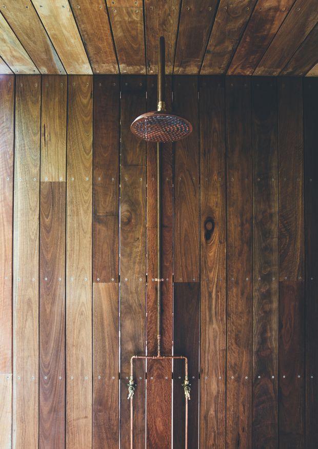 Close-up view of a wall-mounted shower fixture against a full-height dark timber-planked wall and matching timber ceiling. The shower has a round overhead rose, exposed vertical pipework, and lower controls, creating a rustic, materially rich bathroom installation.