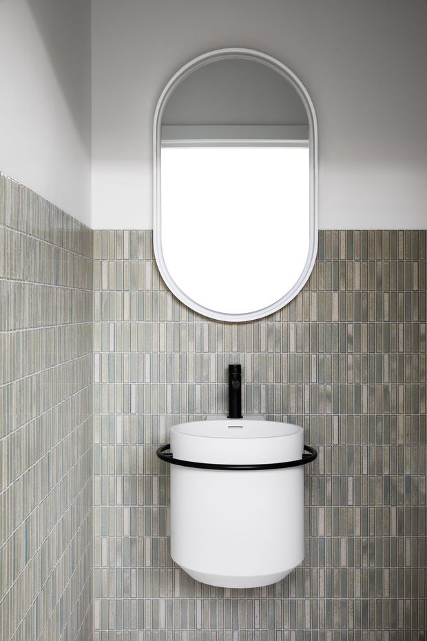 Compact bathroom or powder room featuring a wall-mounted cylindrical white basin with a black circular support rail and black tapware. The walls are clad in vertically arranged narrow rectangular tiles in muted grey-green and beige tones. A large white-framed arched oval mirror is mounted above the basin, creating a clean, graphic focal point.
