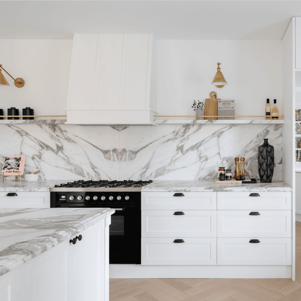 Bright, refined white kitchen with Shaker-style cabinetry, a large veined marble-look splashback and benchtops, black freestanding range cooker, brass-accented open shelving, and a marble-topped island. Herringbone timber flooring and carefully styled accessories add warmth and contrast.