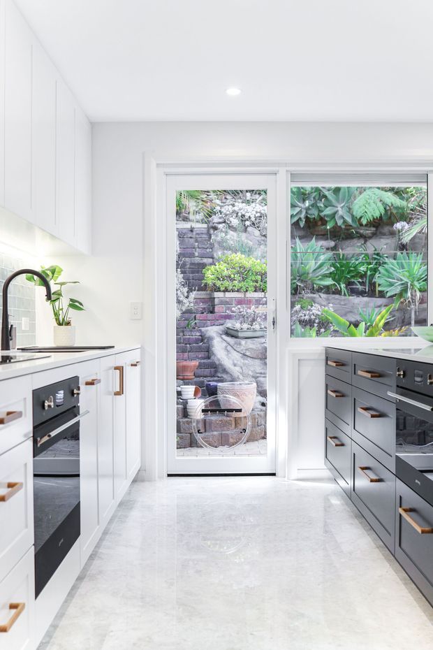 Bright, compact contemporary kitchen with white full-height cabinetry on the left and dark charcoal cabinetry on the right, accented by warm timber handles. A pale polished stone floor and white walls create a clean, luminous setting. A glazed external door and adjacent window overlook a lush planted courtyard and masonry retaining wall. Integrated ovens, a black gooseneck tap, undermount sink, and pale stone benchtops are visible.